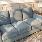 Modern light gray 3-seater fabric sofa with premium upholstery, plush cushions, rounded armrests, and a sturdy wooden base in a contemporary living room.