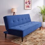 Modern blue fabric sofa cum bed with tufted backrest, wooden legs, and fold-flat sleeper design placed in a contemporary living room with large glass windows and indoor plants.