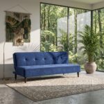 Modern blue fabric sofa cum bed with tufted backrest, wooden legs, and fold-flat sleeper design placed in a contemporary living room with large glass windows and indoor plants.