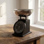 Homslux matte black vintage mechanical kitchen scale with stainless steel bowl on a wooden rustic table next to a bowl of red apples