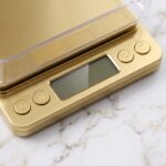Homslux gold digital kitchen scale with a clear plastic weighing tray on a marble countertop next to a bowl of walnuts