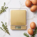Homslux gold digital kitchen scale with a clear plastic weighing tray on a marble countertop next to a bowl of walnuts