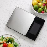 Homslux stainless steel digital kitchen scale with a large black display screen on a white speckled countertop next to fresh vegetables