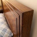 Dark walnut brown single bed with a framed rectangular panel headboard, raised wooden-style legs, cream bedding, knitted beige throw, and neutral bedroom décor.