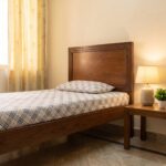 Dark walnut brown single bed with a framed rectangular panel headboard, raised wooden-style legs, cream bedding, knitted beige throw, and neutral bedroom décor.