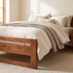 Solid natural wood single bed frame with an open horizontal slatted headboard styled with cozy beige linen bedding and a chunky knit throw blanket.