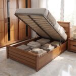 Wooden single bed frame with hydraulic gas-lift mechanism open showing folded blankets and towels stored inside under-bed compartment.